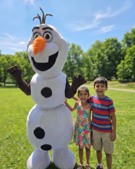 Children posing with a large snowman character in a park on a sunny day.
